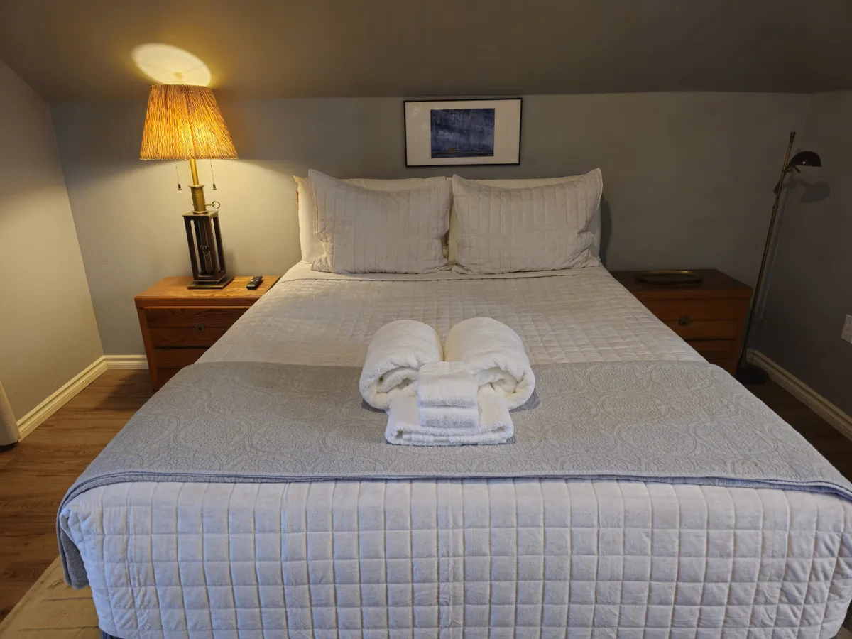 A serene bedroom scene with a neatly made bed, soft lamp lighting, fresh towels, a nightstand, and tasteful artwork. The ambiance is peaceful and inviting, suggesting a space perfect for relaxation and intimacy.