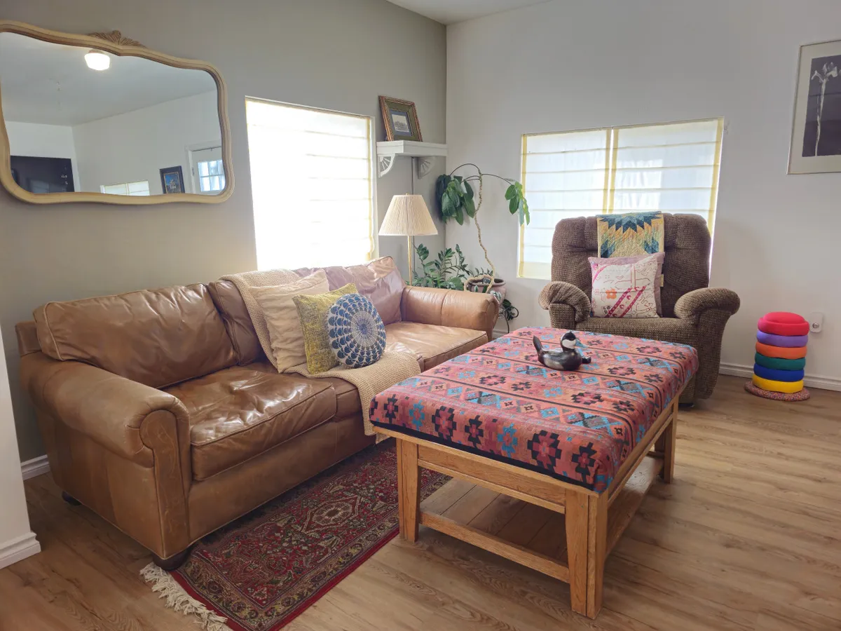 The image showcases a sunlit, inviting living room with a plush leather couch, a vibrant ottoman, leafy potted plant, warm lighting from a stylish lamp, and thoughtfully curated framed artwork. The space exudes comfort and warmth, perfect for unwinding after a day on the slopes or gathering with friends and family for games and conversation.
