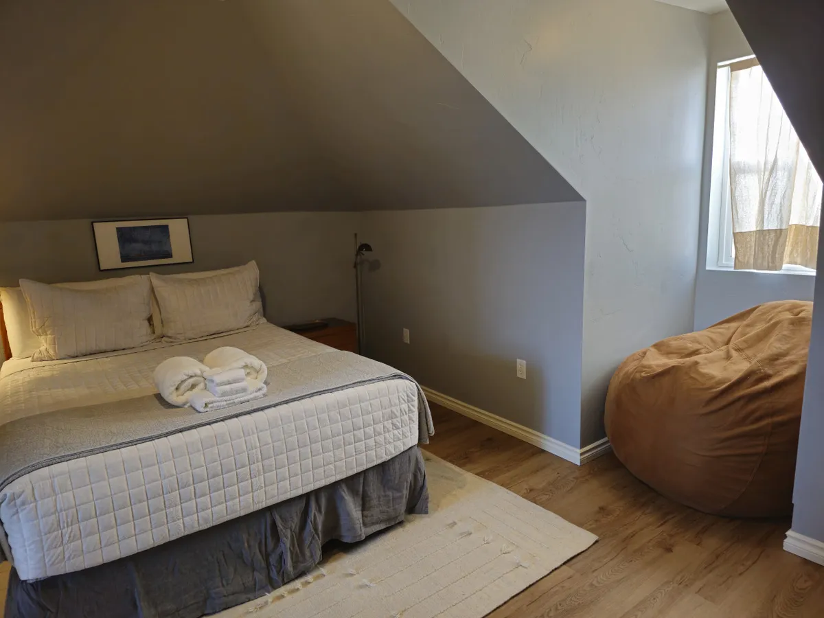 This image features a bed with plush bedding, a bean bag chair, and a window that allows natural light to fill the space. The atmosphere is inviting and restful, suggesting a comfortable place to unwind after a day outdoors.