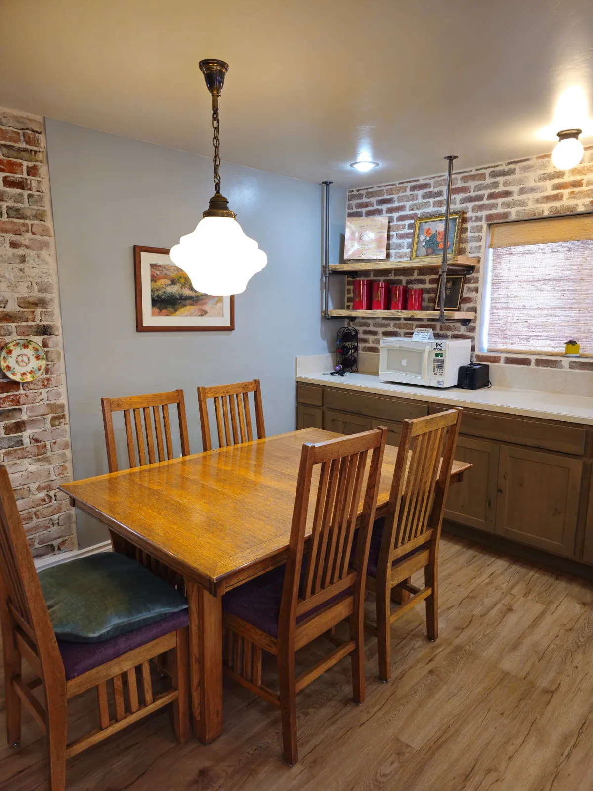 This image features a warm, welcoming dining area showcasing a wooden dining table surrounded by chairs, set against a backdrop of a brick wall and illuminated by a hanging light fixture. The kitchen countertop is visible, hinting at the connection between meal prep and dining. The overall atmosphere reflects active family life, with space for shared meals, conversations, and gathering.