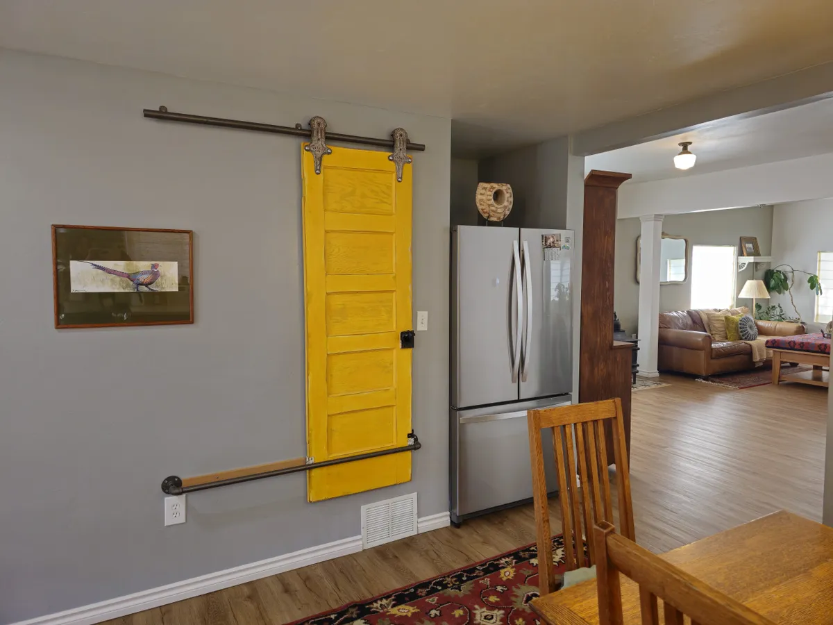 The image captures a homey and inviting kitchen-dining space with a stainless steel refrigerator, a wooden dining table, a yellow sliding door, and framed bird art. The setting conveys a sense of everyday family life, blending modern appliances with rustic, personalized touches.