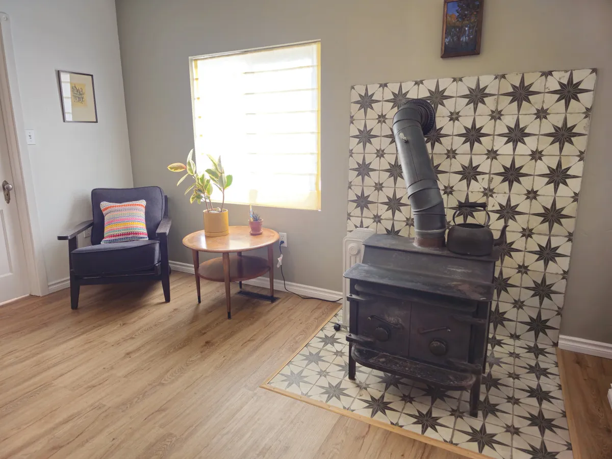 A welcoming living area featuring a wood stove framed by eye-catching decorative tiles. An armchair and coffee table invite guests to gather, while natural light streams in through a window, emphasizing the cozy, communal vibe of the space.