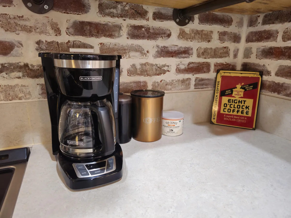 A warmly lit kitchen corner featuring a coffee maker, coffee can, and storage containers. The space suggests morning rituals, casual conversations, and the simple pleasure of sharing a fresh cup of coffee together.
