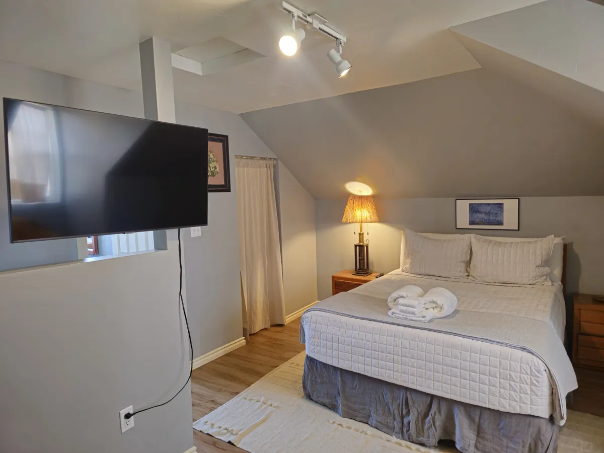A serene bedroom scene featuring a neatly made bed with plush pillows and fresh towels, a bedside lamp casting a gentle glow, and a wall-mounted television. The decor is soothing and uncluttered, embodying tranquility and comfort ideal for restful nights.